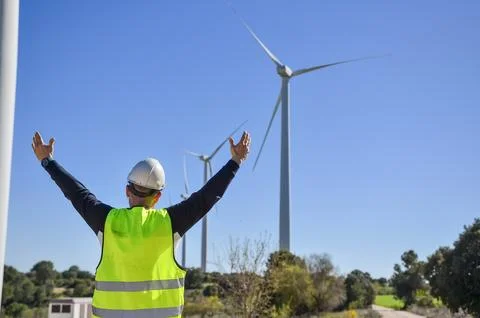 Engineer raising arms in front of wind turbines, celebrating renewable energy Stock Photos