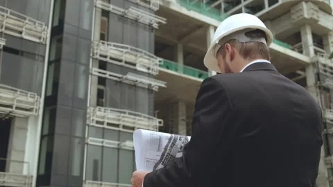 Engineer in a suit and white safety helmet standing with blueprint at Stock Footage 313359679