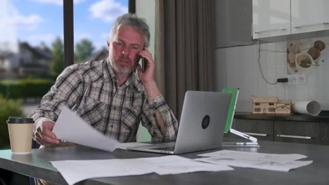 Engineer talking on phone while reviewing technical paperwork at kitchen table Stock Footage 334870965