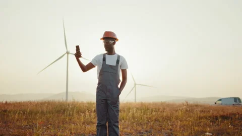 Engineer in uniform using modern smartphone for video call among windmill farm Stock Footage 170660551