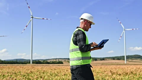 Engineer uses a tablet in a field beside wind turbines during daytime Stock Footage 348698562