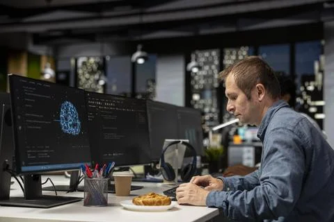 Engineer utilizing AI on computer to optimize performance while eating croissant Stockfoto's