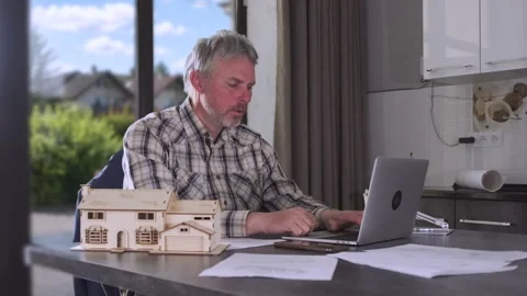 Engineer working on laptop at kitchen table with house model and technical Stock Footage 334870963
