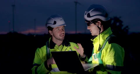 Engineers in safety uniform working at night near wind turbines using laptop and Stock Footage 335210095