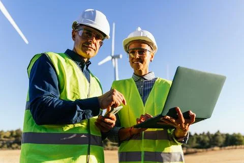 Engineers using laptops to communicate while standing in front of wind turbines. Stock Photos