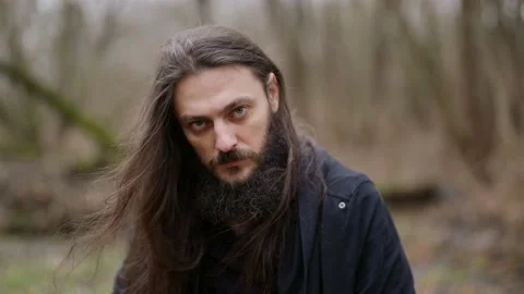 Enigmatic man with beard and long hair is wearing black clothes, looking at Video stock 167680648