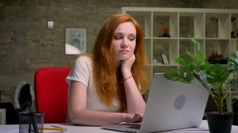 Enjouable cute thoughtful red-haired caucasina woman is smiling at laptop and Video stock 98301636