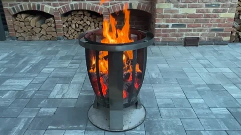 Enjoying fire in a metal fire pit on a patio with bricks and stacked wood in the Stock Footage 339574903