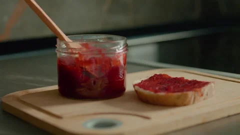 Enjoying a slice of bread topped with raspberry jam on a kitchen chopping board Stock Footage 320952031