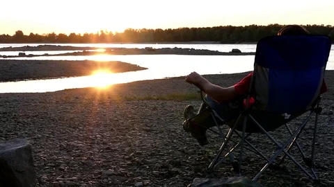 Enjoying the sunset sitting on a stool on a stony bank near the river at sunset Stock Footage 80987193