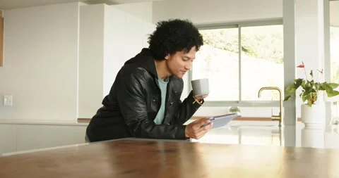 Entering bright kitchen, man sipping from gray mug while browsing tablet at Stock Footage 345035496