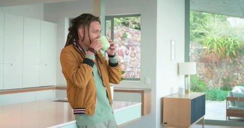 Entering kitchen, 30s man with dreadlocks leaning on island sipping green mug Stock Footage 336242711