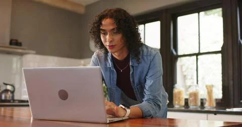 Entering kitchen, non-binary person placing laptop on wooden island and typin Stock Photos