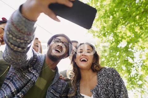 Enthusiastic friends taking selfie with camera phone below tree Stock Photos