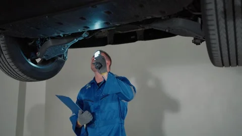 EV Mechanic inspects vehicle under lift with flashlight and clipboard Video stock 338332406