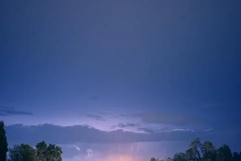 Evening blue sky with lightning sparkling in the clouds above the trees Stock Photos