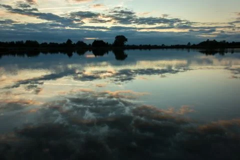 Evening clouds in the blue sky reflecting in the water of a calm lake Stock Photos