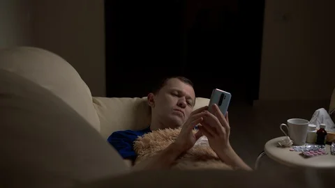 In the evening, a man lies alone on the couch, is sick, uses his phone Stock Footage 129759494