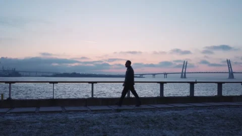 Evening walker moves swiftly on seaside boardwalk, iconic bridge spanning Stock Footage 271297923