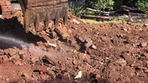 Excavator bucket scooping up rubble, worker splashing water to keep down dust.  Video stock 252343898