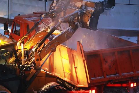 An excavator loads snow into the back of a dump truck at night. Stock-Fotos