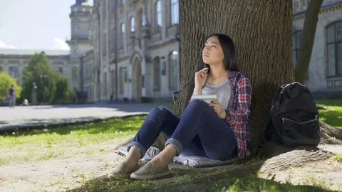 Exchange student sitting under tree, holding notebook, thinking, check-in list 動画素材 77199417
