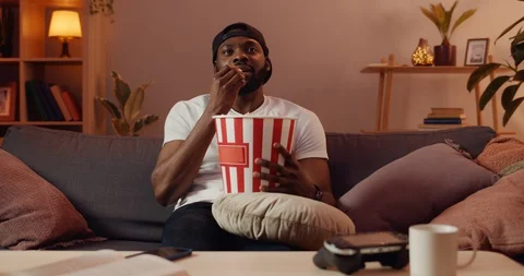 Excited guy eating popcorn from paper bowl and smiling while looking at TV