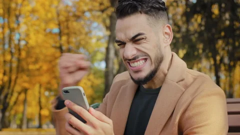 Excited hispanic guy reading good news on smartphone sitting on bench in autumn 스톡 동영상 165826738
