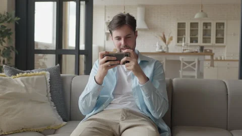 Excited Man Playing Mobile Game On Sofa Fully Engaged In Competitive Gaming Video stock 302450334