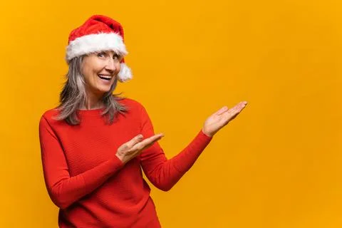 Excited middle-aged gray-haired woman in red santa hat standing isolated on Stock Photos