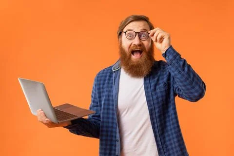 Excited redhaired bearded programmer guy holds laptop over orange background 스톡 사진