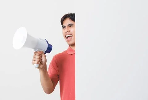 Excited young man shouting through a megaphone while peeking from behind a .. Stock Photos