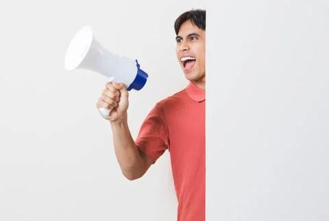 Excited young man shouting through a megaphone while peeking from behind a .. Stock Photos