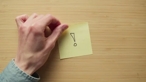Exclamation mark drawn on post it note is placed onto a bamboo desk top Stock Footage 320557699