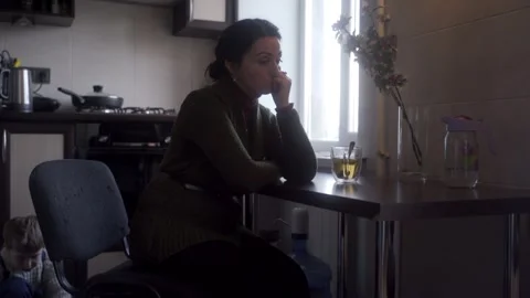 Exhausted mother sits at kitchen table, looking tired with young child playing Vidéo 273513770