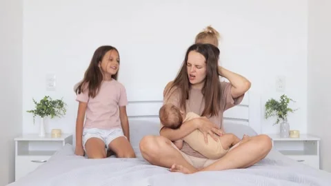 Exhausted mother sitting on bed with hand on head feeling migraine while caring Stock Footage 291267757