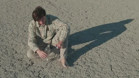 An exhausted young man sits on the lifeless ground under the rays of the Stock Footage 253707059
