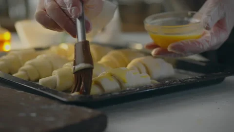 An experienced baker wearing protective gloves brushes croissants in an indu Stock Footage 323708724