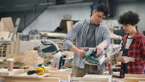 Experienced joiner teaching female apprentice to use belt sander polishing wood Stock Footage 113737184