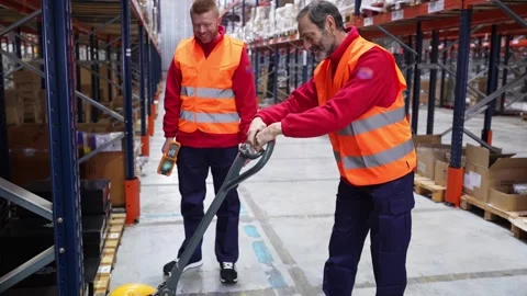 Experienced warehouse worker showing new employee how to use pallet jack Stock Footage 312856352
