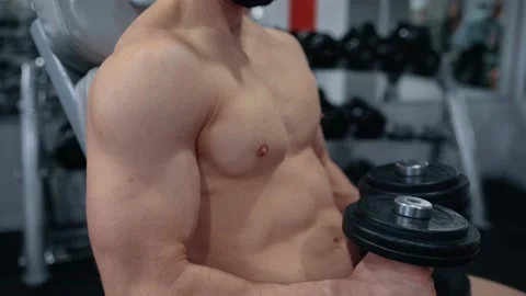 Exploring the young bodybuilder sitting on a chair with dumbbells in his hand Stock Footage 156477632