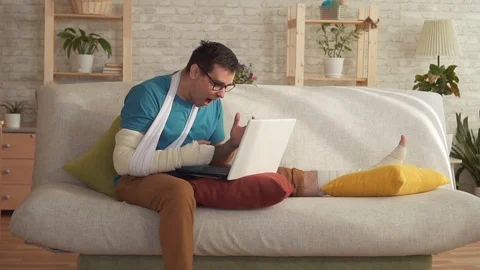 Expressive young man with a broken arm and leg uses a laptop and learns about Stock-Footage 106594720