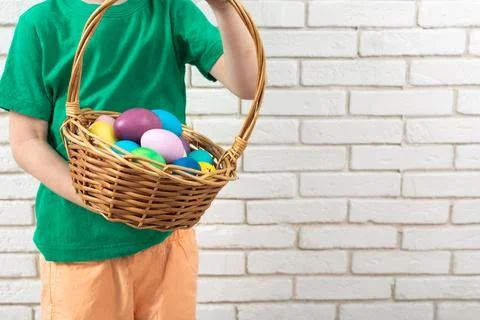 Faceless boy with colorful Easter eggs in wicker basket near white brick wall 스톡 사진