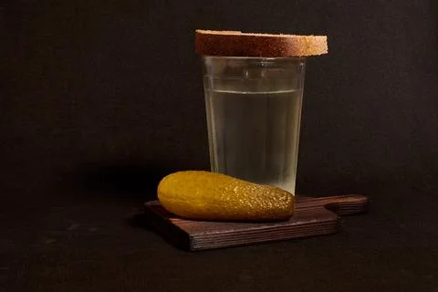 A faceted glass of vodka, rye bread and a pickled cucumber on a wooden stand Stock Photos