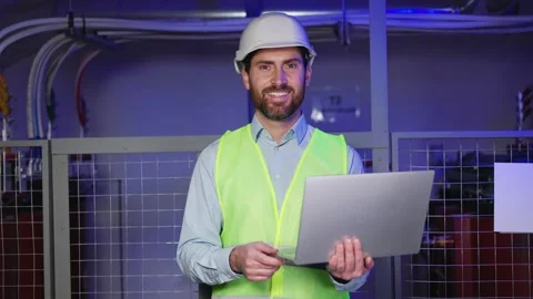 Factory worker in hard hat, green vest using laptop, serious electrician at work 動画素材 256138165