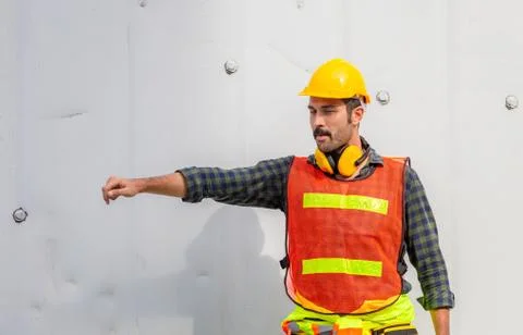 Factory worker man in hard hat and safety vest extends a hand at containers c Фото