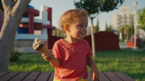 Fair-haired boy eats white cold cream ice cream in waffle cone cup while sitting Video stock 201657874