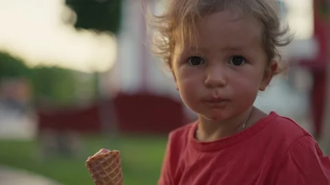 Fair-haired boy eats white cold cream ice cream in waffle cone cup while sitting Vidéo 201964803