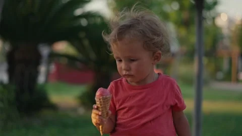 Fair-haired boy eats white cold cream ice cream in waffle cone cup while sitting Stockbeeldmateriaal 202175485