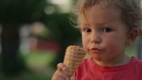 Fair-haired boy eats white cold cream ice cream in waffle cone cup while sitting 스톡 동영상 203790652
